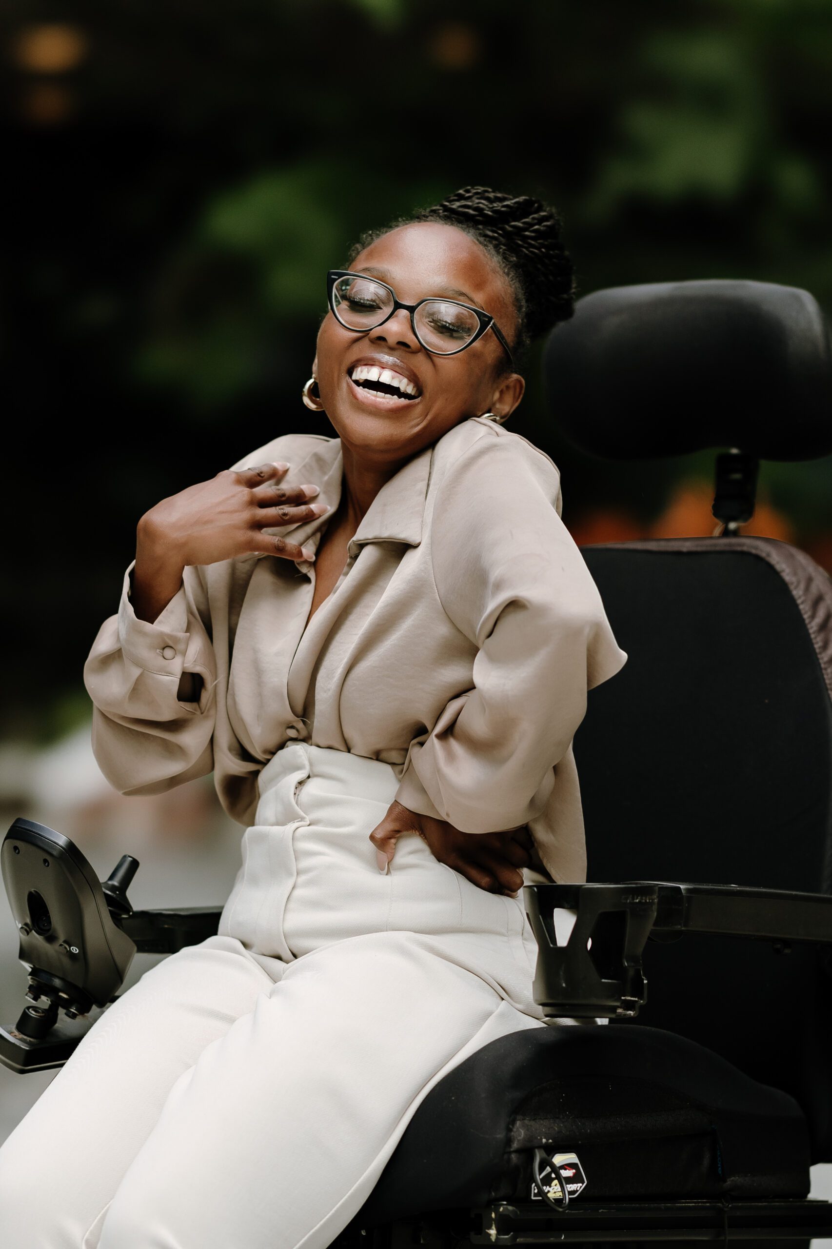 A portrait of a person wearing glasses, a tan jacket and white pants sitting in in an electric wheelchair and laughing
